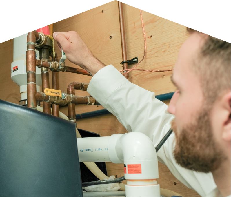 Peel water heater specialist tightens a valve on copper piping during a water heater installation, with exposed wood paneling and wiring in the background.